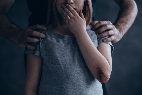 man with his hands gripping the child's shoulders as the child holds her hand over her mouth in fear, against a grey background